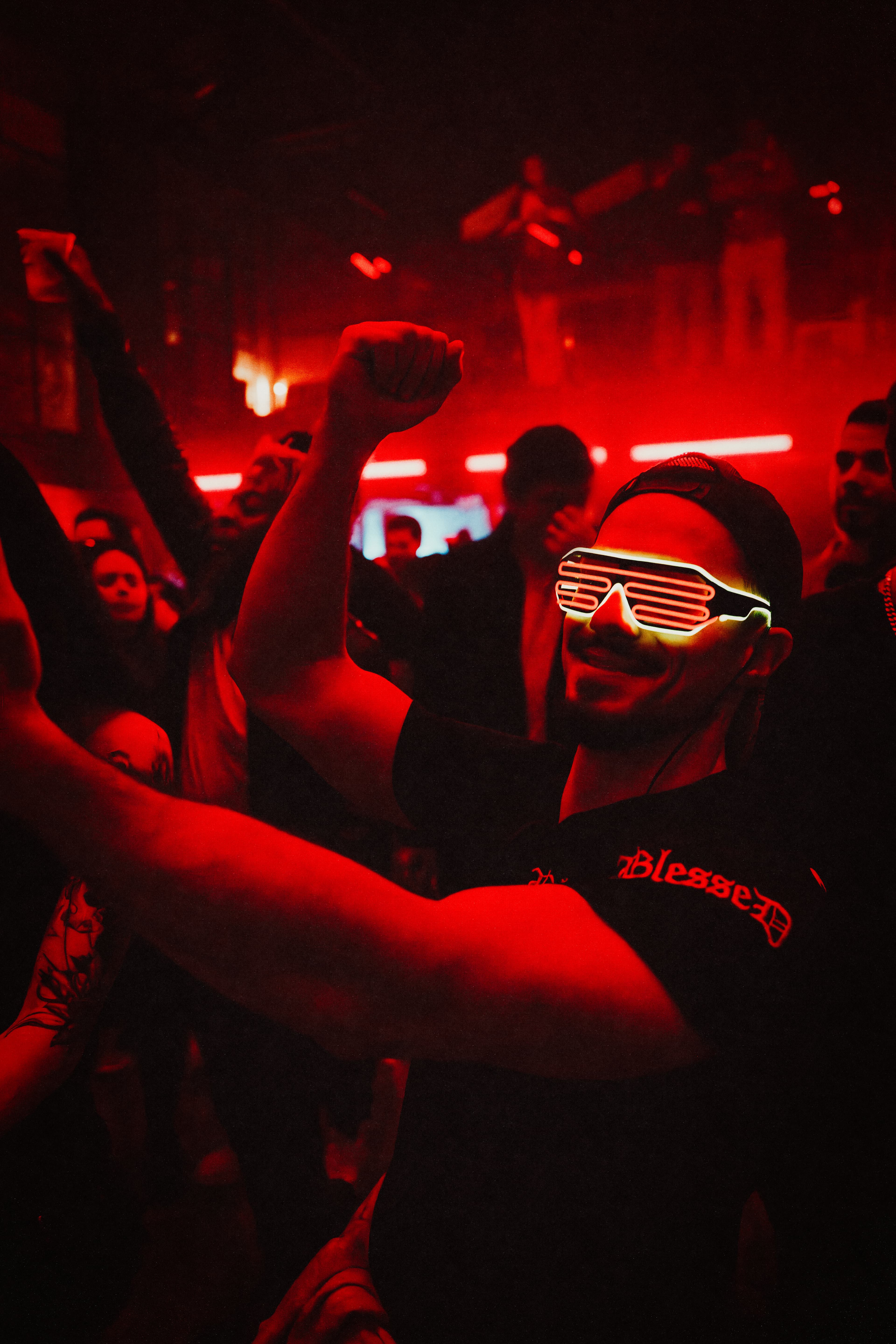 Man wearing glowing LED glasses cheers with a raised fist in a vibrant red nightclub.