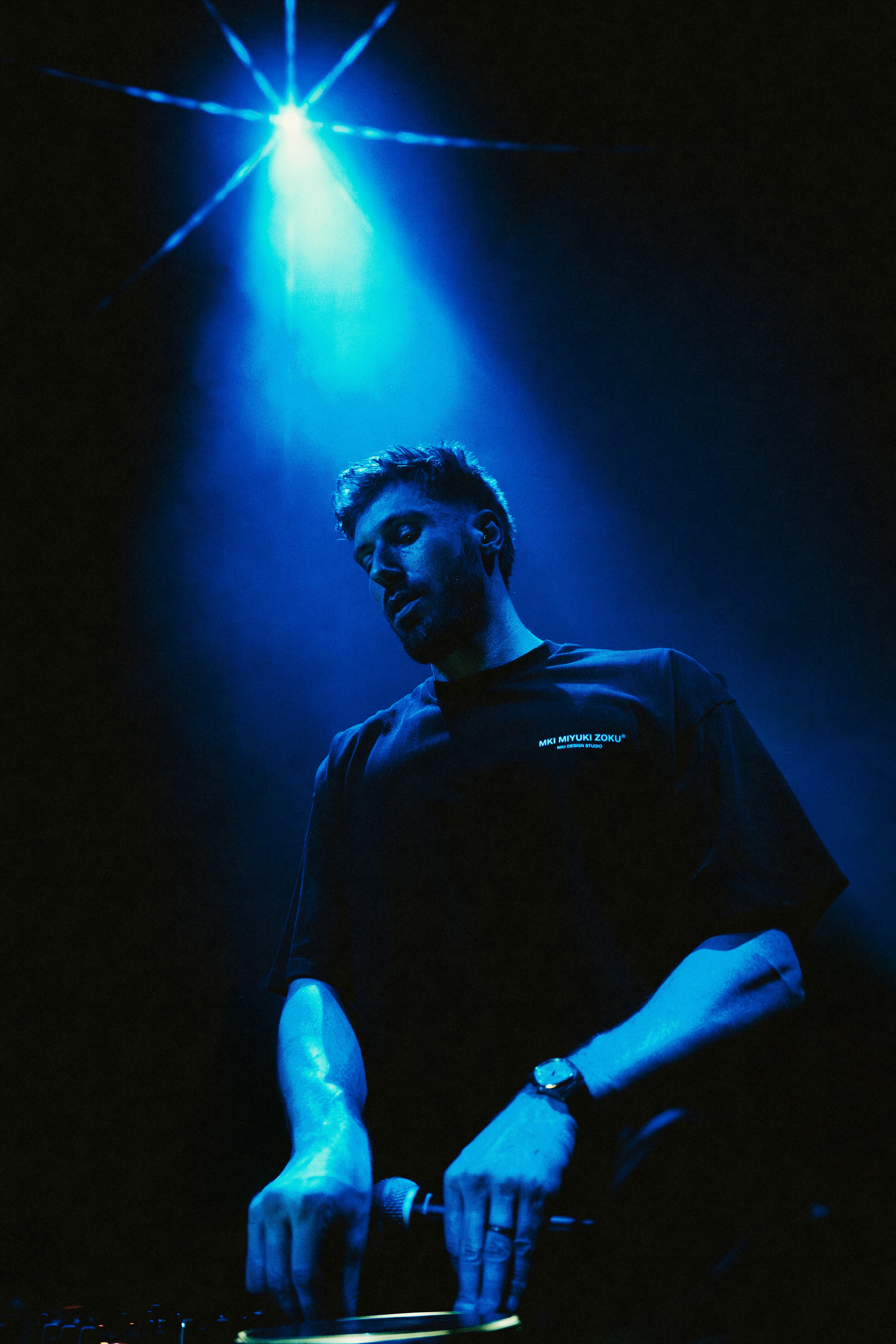 A DJ performs on a dark stage, illuminated by a single, intense blue spotlight.