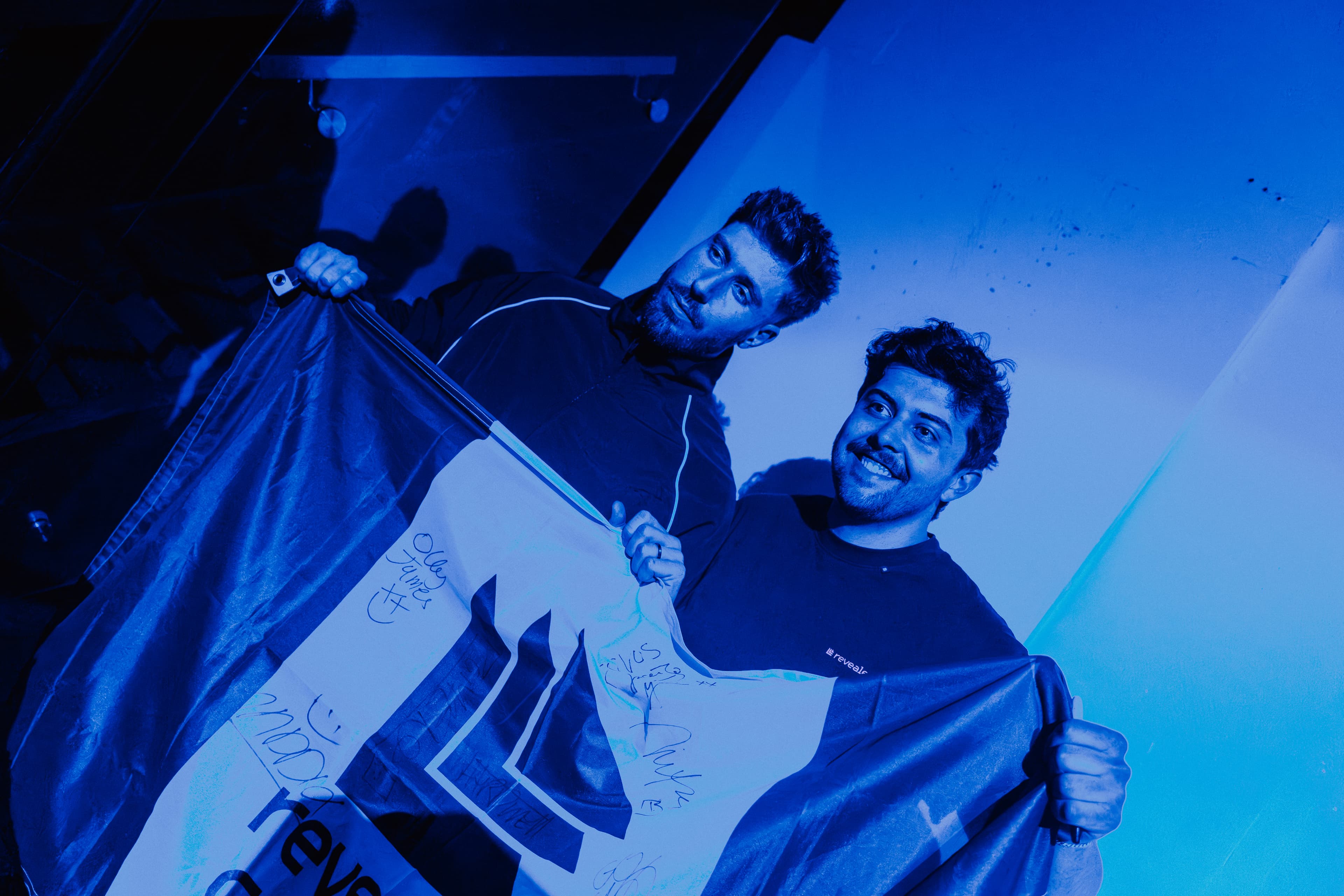 Two men hold a signed white banner with a logo under vibrant blue lighting.